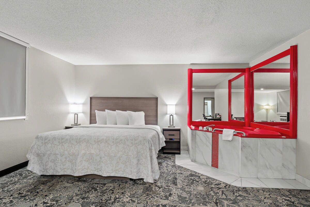 Habitación de hotel con cama, mesitas de noche, baño con lavabo y espejo rojos, y una alfombra estampada.