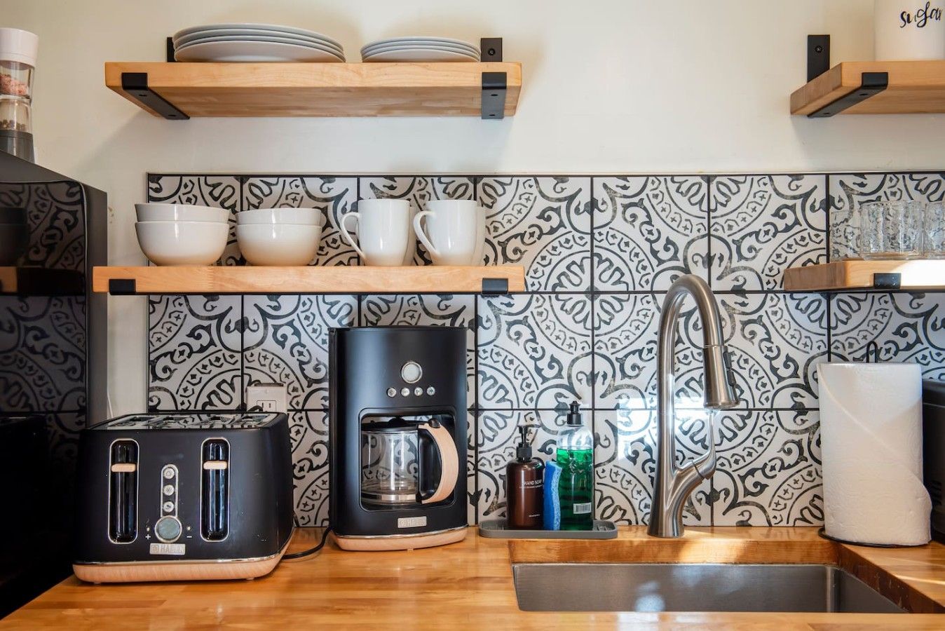 A kitchen with a toaster, coffee maker, and a sink. There are shelves with dishes and a patterned backsplash.