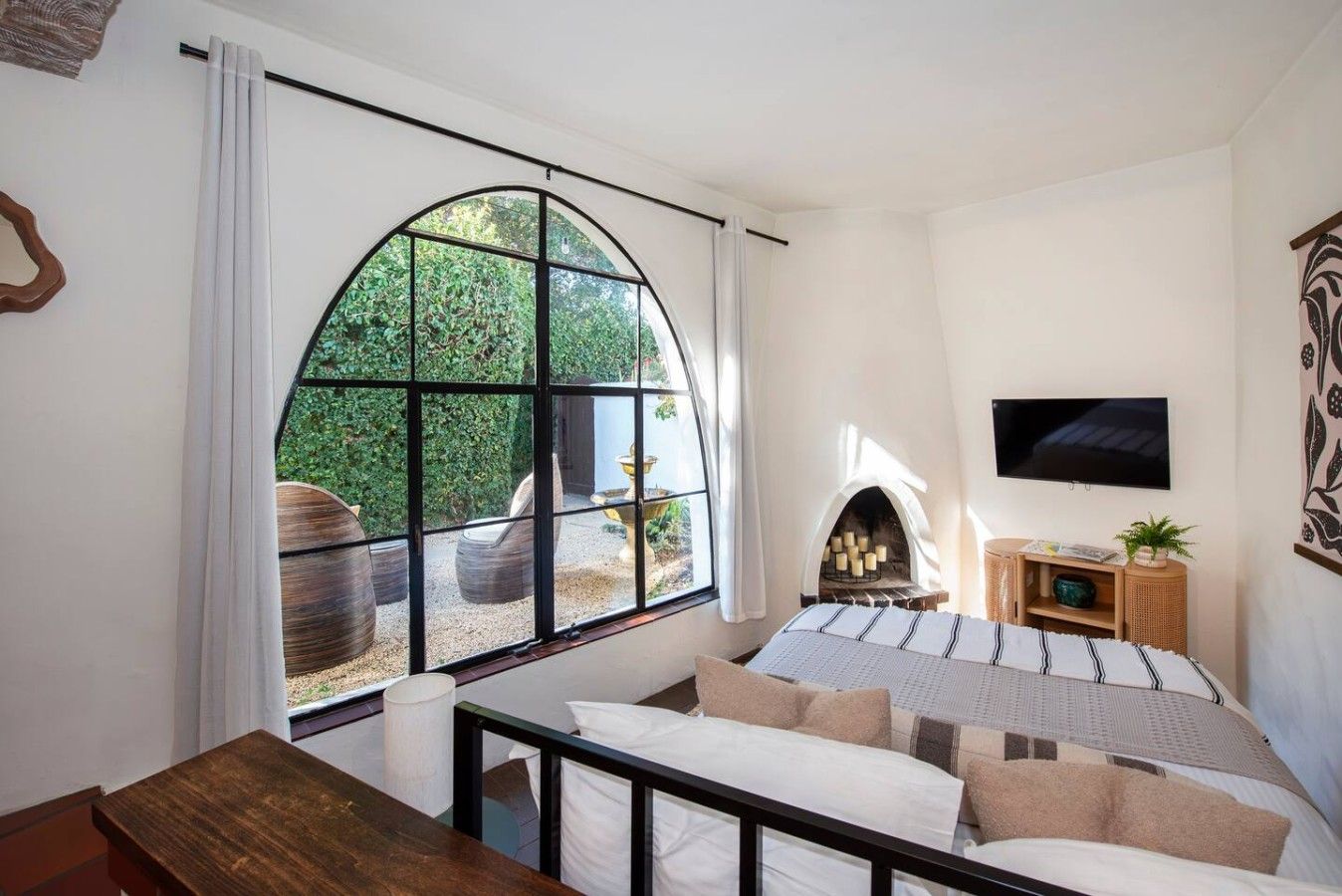 A bedroom with a large arched window, a fireplace, a television, and a wooden bench.