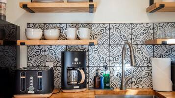 A kitchen with a toaster, coffee maker, and a sink. There are shelves with dishes and a patterned backsplash.