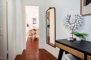 A hallway with a tiled floor, a wooden chair, a table with a vase, a framed picture, a mirror, and a decorative wall piece. - Montecito Studio w/ Patio & Outdoor Charm (Montecito)