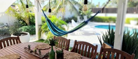 A wooden table with woven placemats, a green bottle, and small plants. A hammock is suspended above, and a pool is visible in the background.