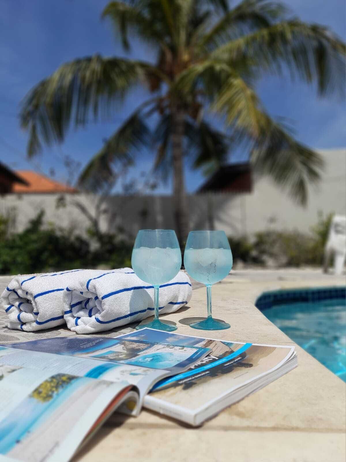 A poolside setting with two glasses of a clear beverage, a folded towel, and a magazine on a table.