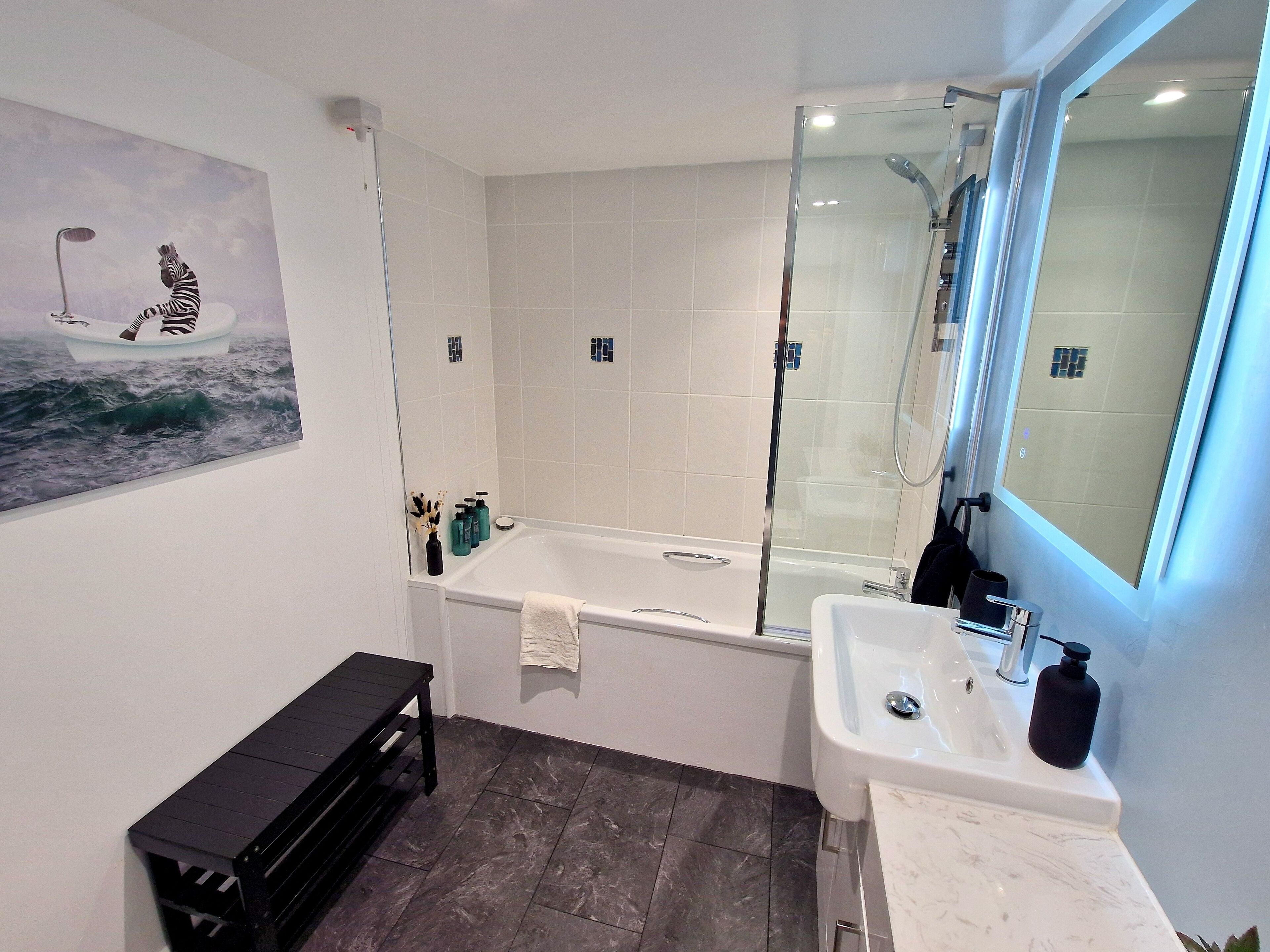 Een moderne badkamer met een badkuip, douche en wastafel. Er is een bankje en een schilderij aan de muur.