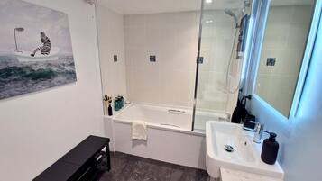 Een moderne badkamer met een badkuip, douche en wastafel. Er is een bankje en een schilderij aan de muur.
