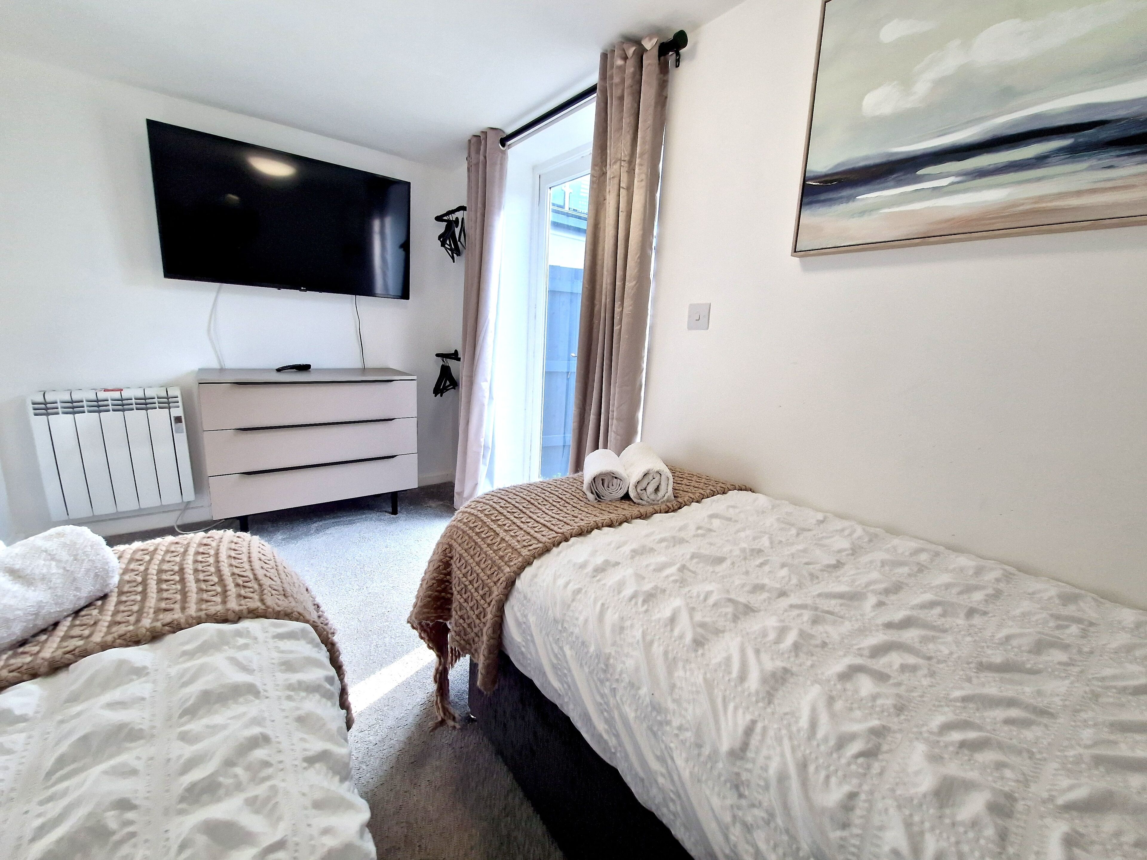 Une chambre d’hôtel avec deux lits, une télévision, une commode, un radiateur et une fenêtre avec des rideaux.