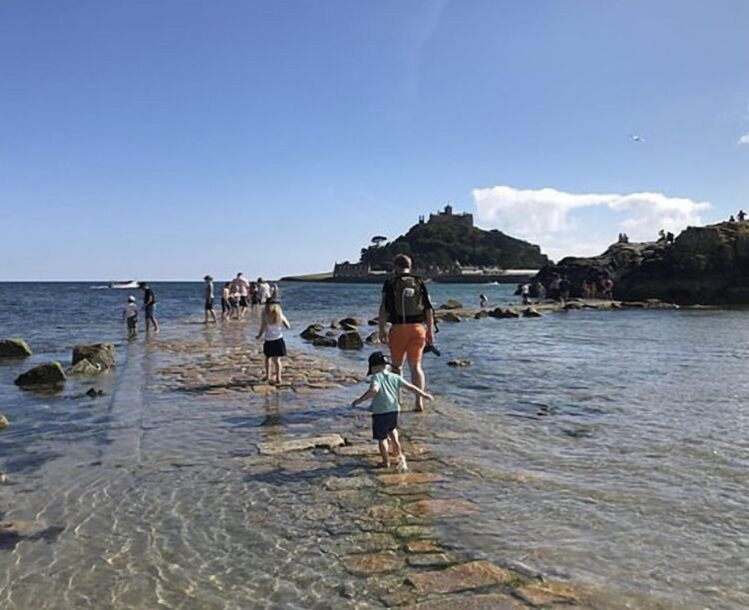 Un rivage rocheux bordé d’une eau claire, où l’on voit des gens se promener, et une structure ressemblant à un château en arrière-plan.