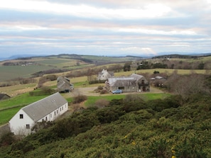 A rural landscape with a white building, a stone barn, and a blue car parked in front.