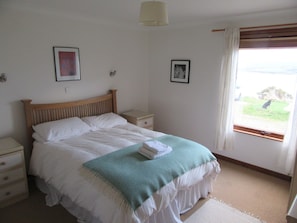 Quarto com cama, mesas de cabeceira, janela com cortinas e duas molduras na parede.