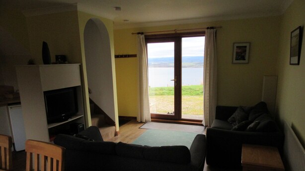 A living room with a sofa, a television, a wooden table, and a view of a body of water through a door. - Osprey, Longhouse Cottages (Fortrose)