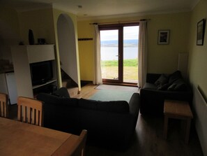 A living room with a sofa, a television, a wooden table, and a view of a body of water through a door. - Osprey, Longhouse Cottages (Fortrose)