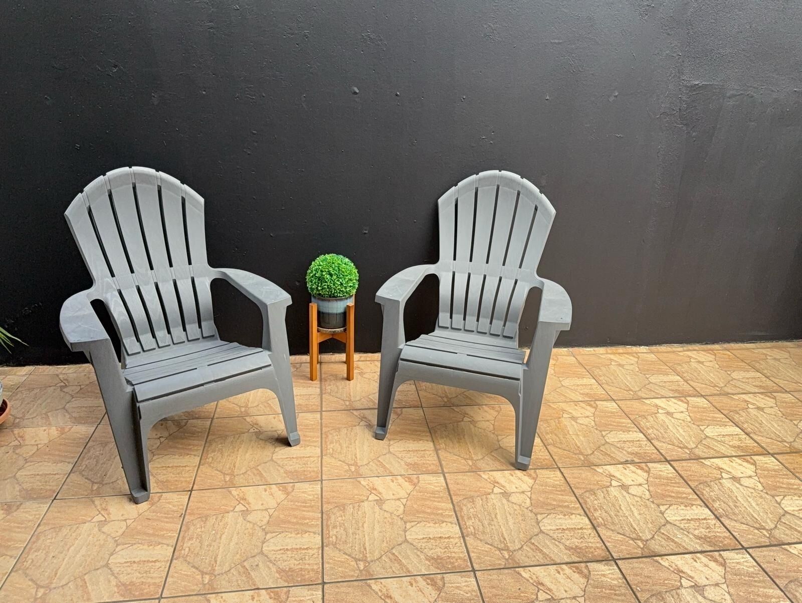 Deux chaises en plastique gris, un petit tabouret en bois et une plante en pot placés entre elles, le tout adossé à un mur noir sur un sol carrelé.