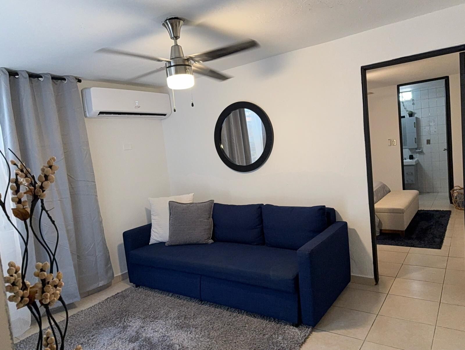 Un salon avec un canapé bleu, un ventilateur de plafond, un miroir rond et une vue sur la salle de bain.