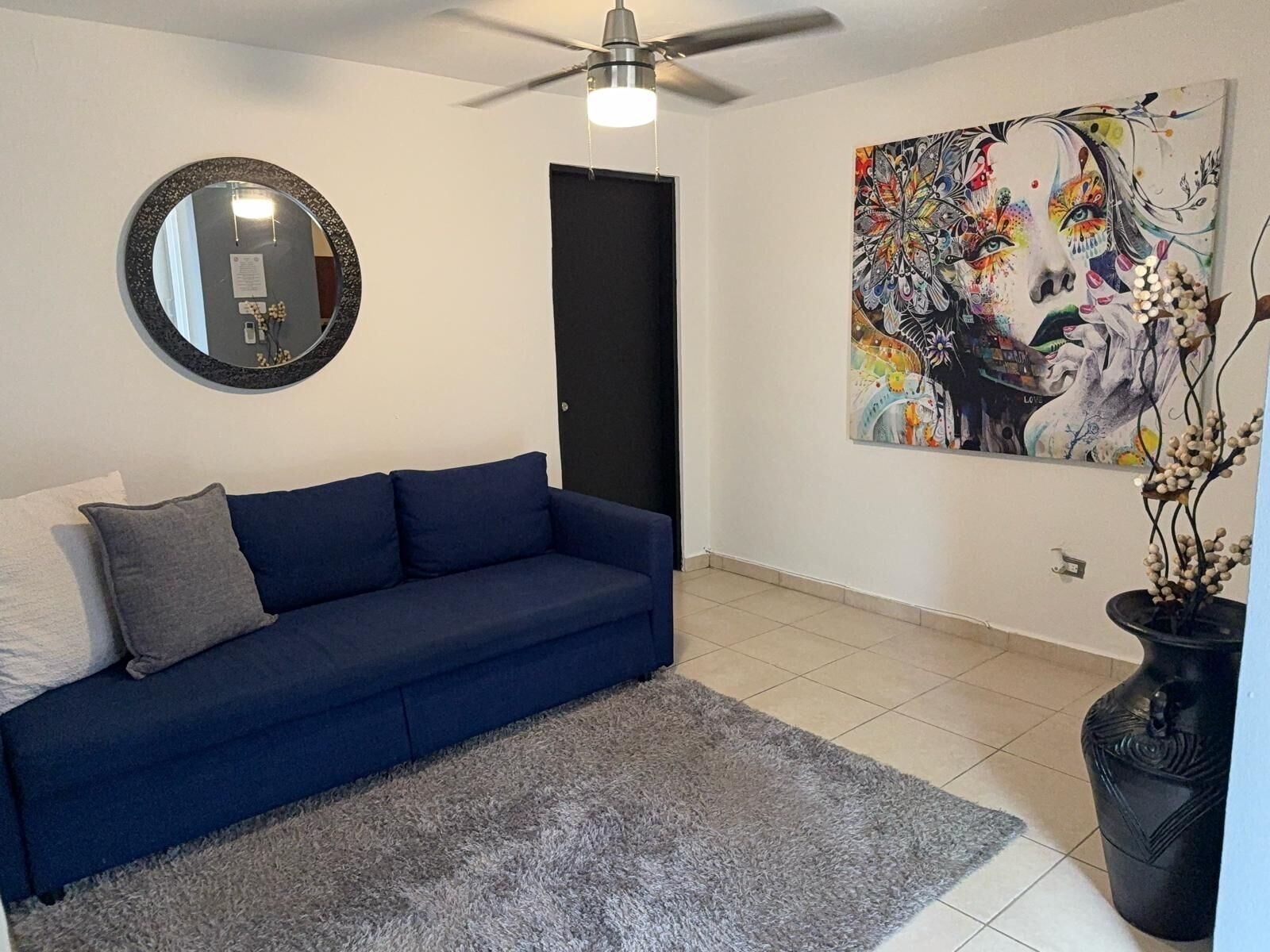 Un soggiorno con un divano blu, uno specchio rotondo, una ventola a soffitto, un quadro astratto di grandi dimensioni e una pianta in vaso.