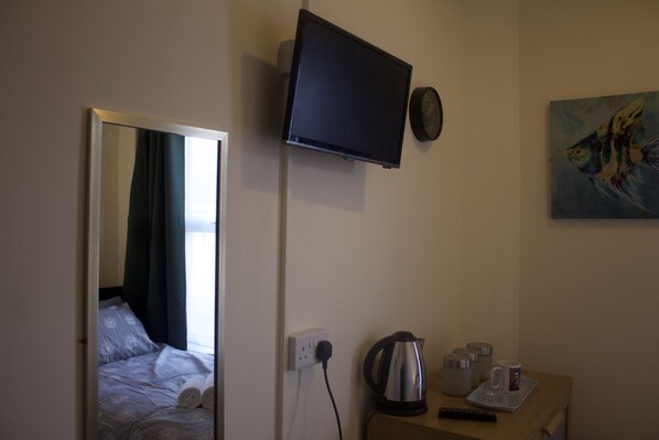 Una habitación de hotel con una cama, un televisor de pantalla plana montado en la pared, un cuadro, un hervidor y una taza sobre una mesa.