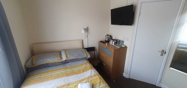 A small, simple room with a bed, a wooden dresser, a television on the wall, and a door. - Elm Tree Guest House (Weston-super-Mare)