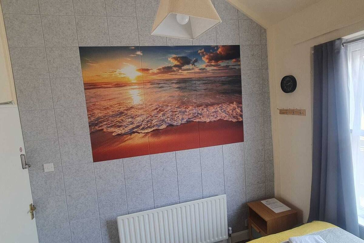 A room with a large painting of a sunset over the ocean, a radiator, a clock, and a window with curtains.
