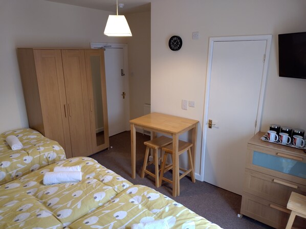 Quarto com cama, guarda-roupa de madeira, mesa pequena com cadeira, relógio na parede e televisão na parede.