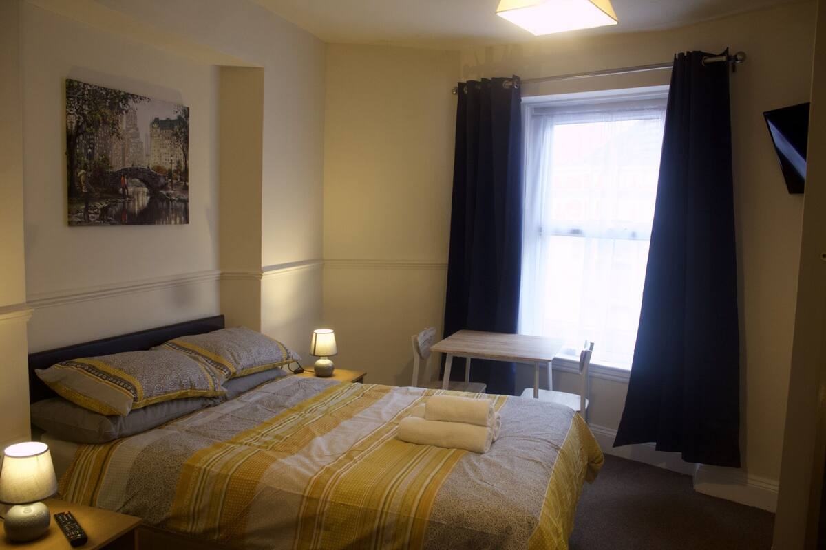 A hotel room with a bed, bedside lamps, a table, a chair, and a window with curtains.