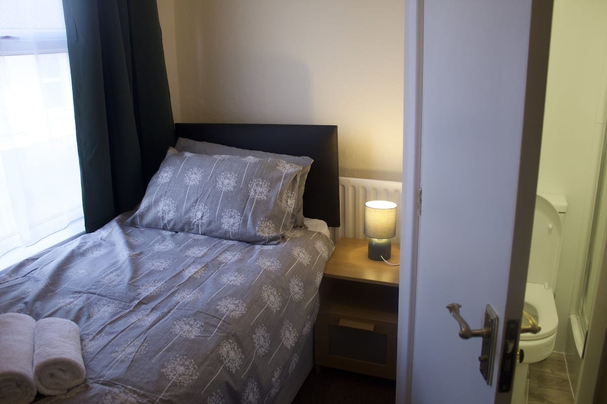 A neatly made bed with a patterned bedspread, a bedside lamp, and rolled towels on the side.