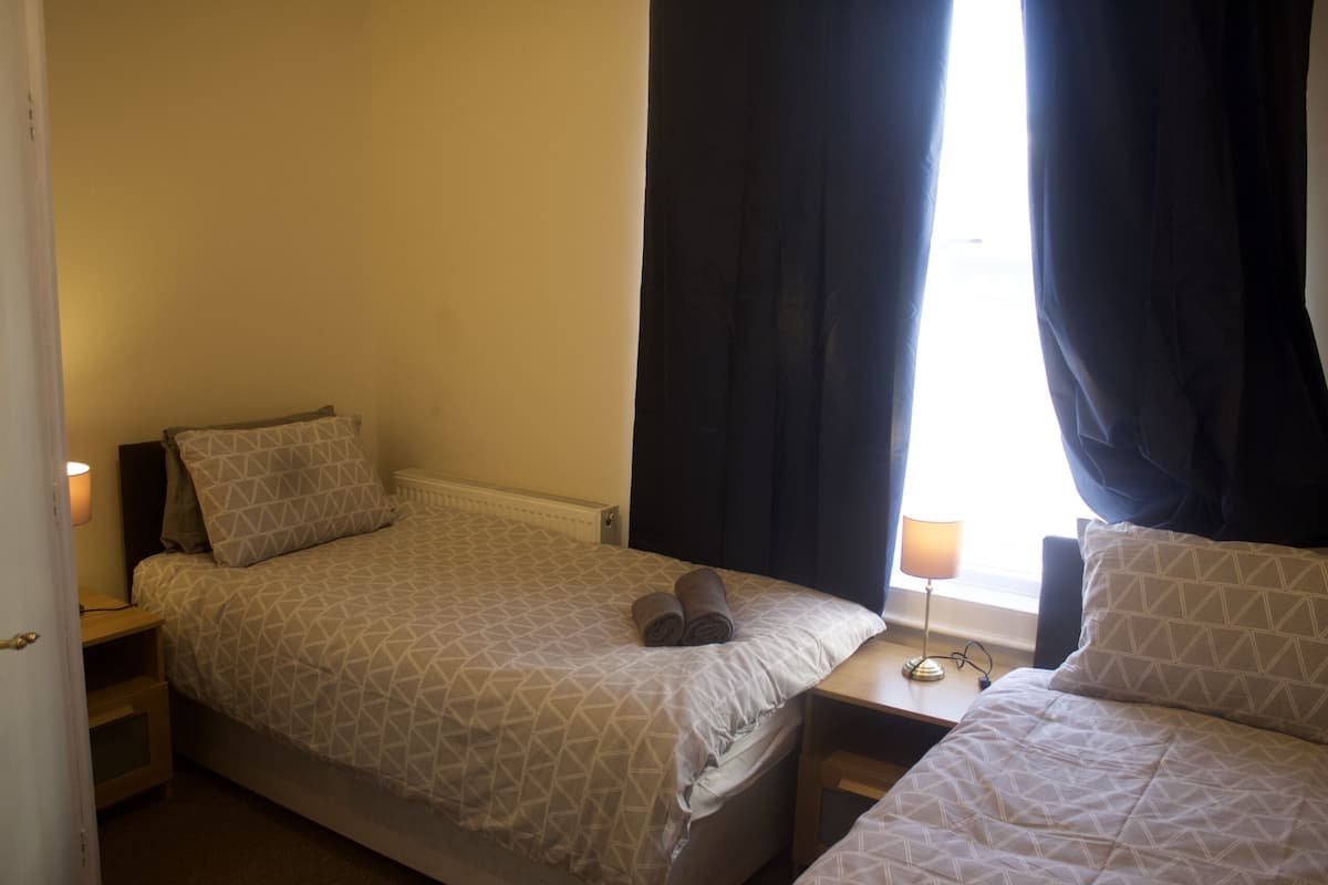 A bedroom with two beds, a bedside table with a lamp, and a window with curtains.