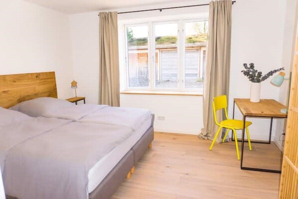 Un dormitorio con cama, escritorio, dos sillas, ventana con cortinas y una mesita de noche con lámpara.
