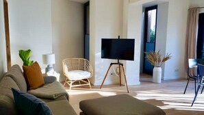 Ein modernes Wohnzimmer mit einer Couch, einem Sessel, einem Fernseher auf einem Stativ und einem Essbereich mit Tisch und Stühlen.