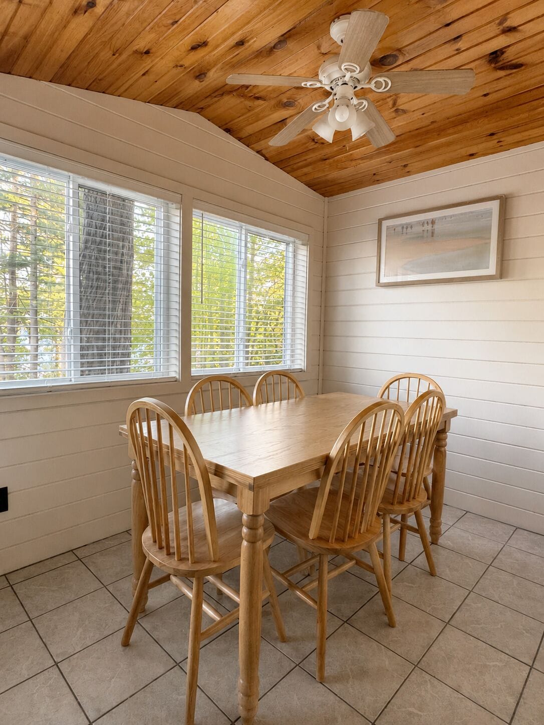 Une salle à manger avec une table et des chaises en bois, un ventilateur de plafond et des fenêtres équipées de stores.