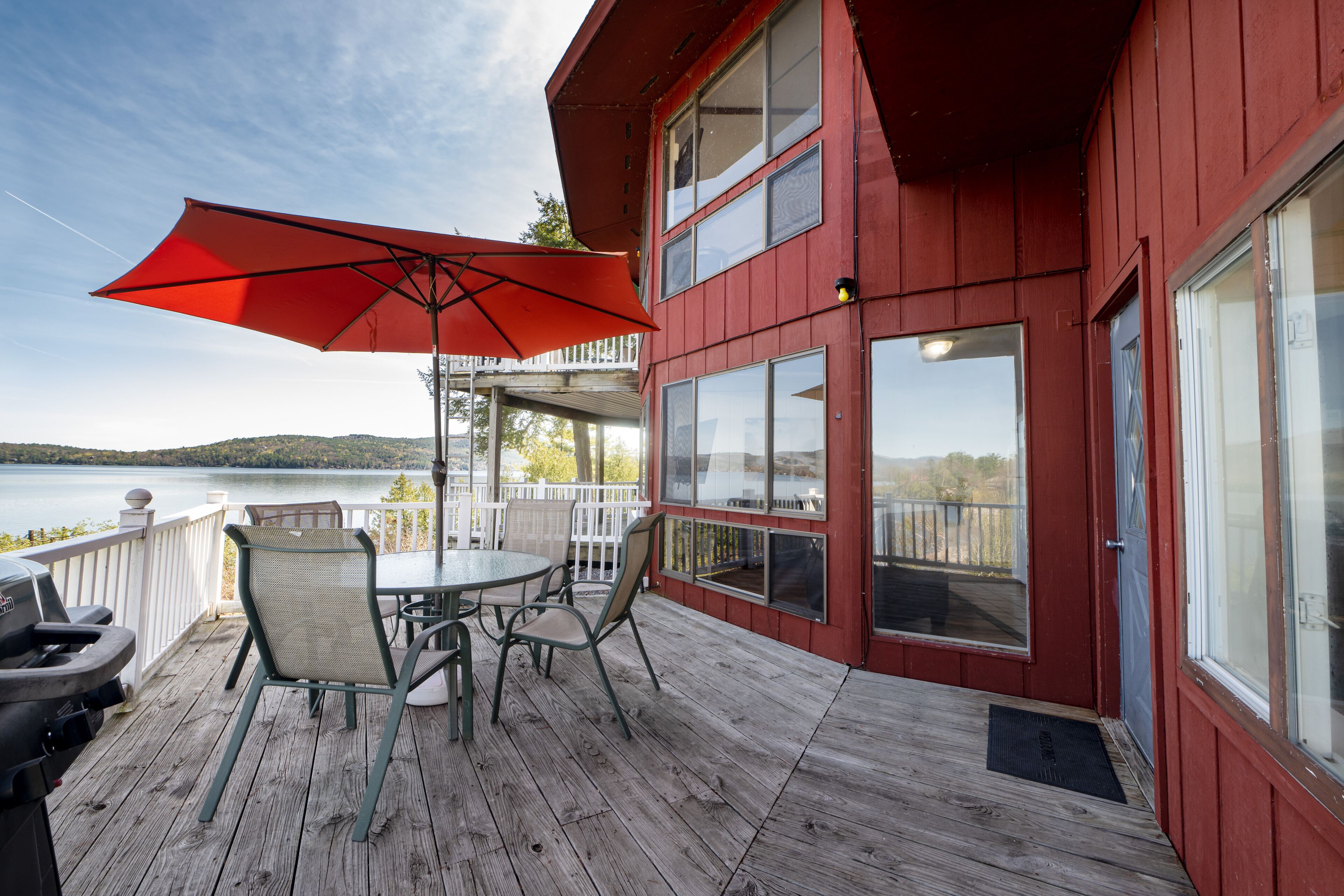 A wooden deck with a table and chairs, a red umbrella, and a view of a lake.