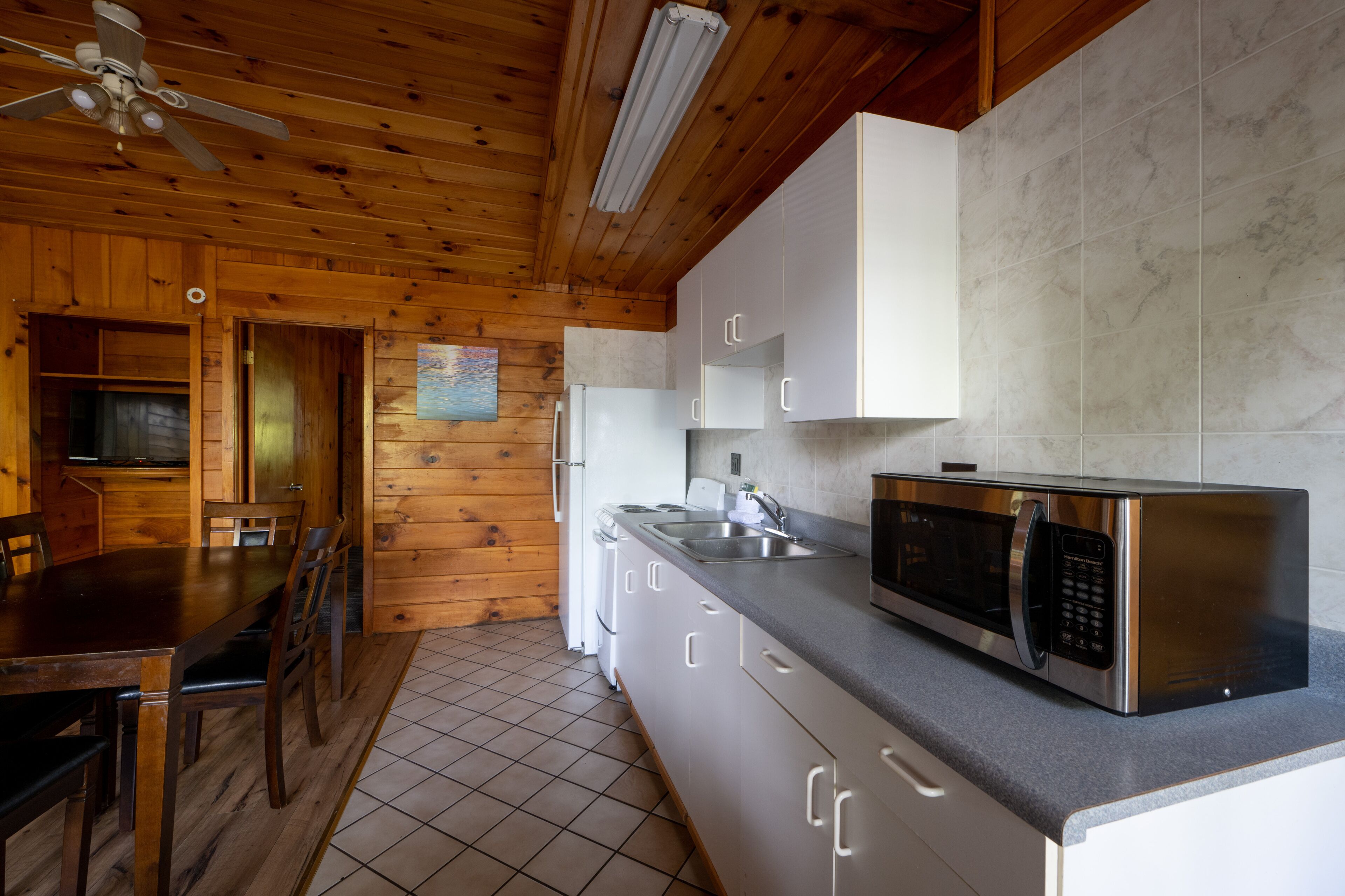 A wooden interior with a kitchen featuring a stainless steel microwave, white cabinets, and a tiled floor.