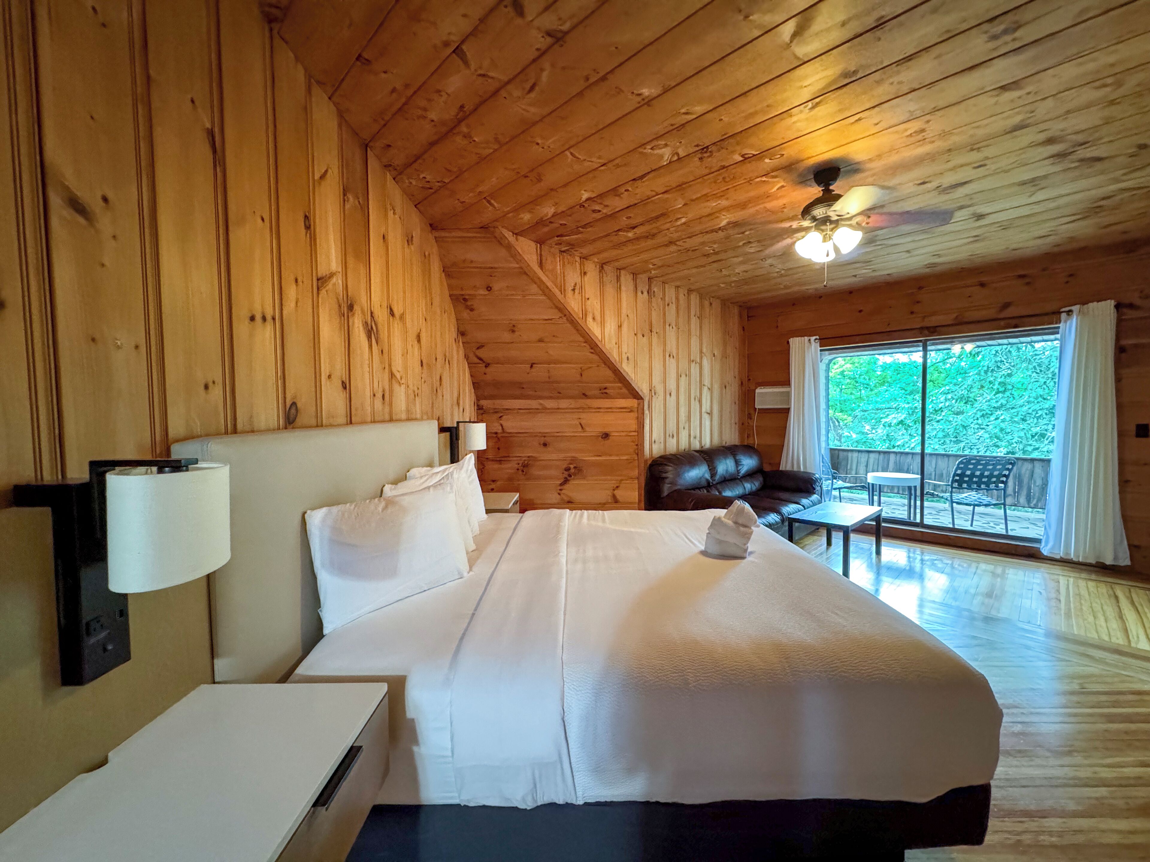 Quarto de cabana de madeira com uma cama, uma mesa de cabeceira, um ventilador de teto e vista do exterior por uma grande janela.