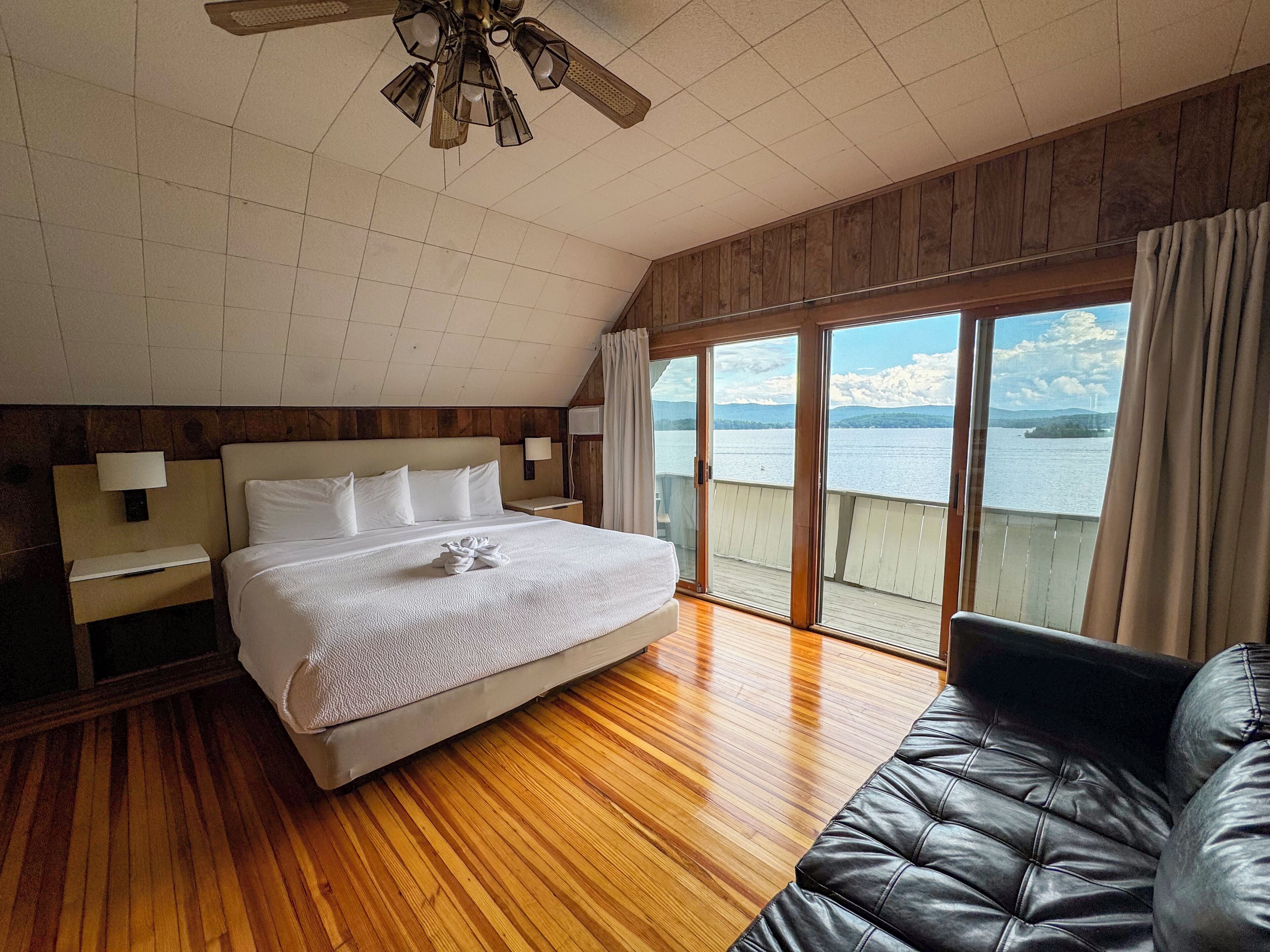 A bedroom with a large bed, wooden flooring, a ceiling fan, a nightstand, a lamp, a sofa, and a view of a lake through large windows.