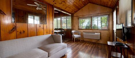 A wooden cabin interior with a sofa, a ceiling fan, a TV, and a window with blinds.