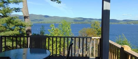 Une terrasse en bois avec une table et des chaises en verre, donnant sur un lac et offrant une vue sur des montagnes au loin.
