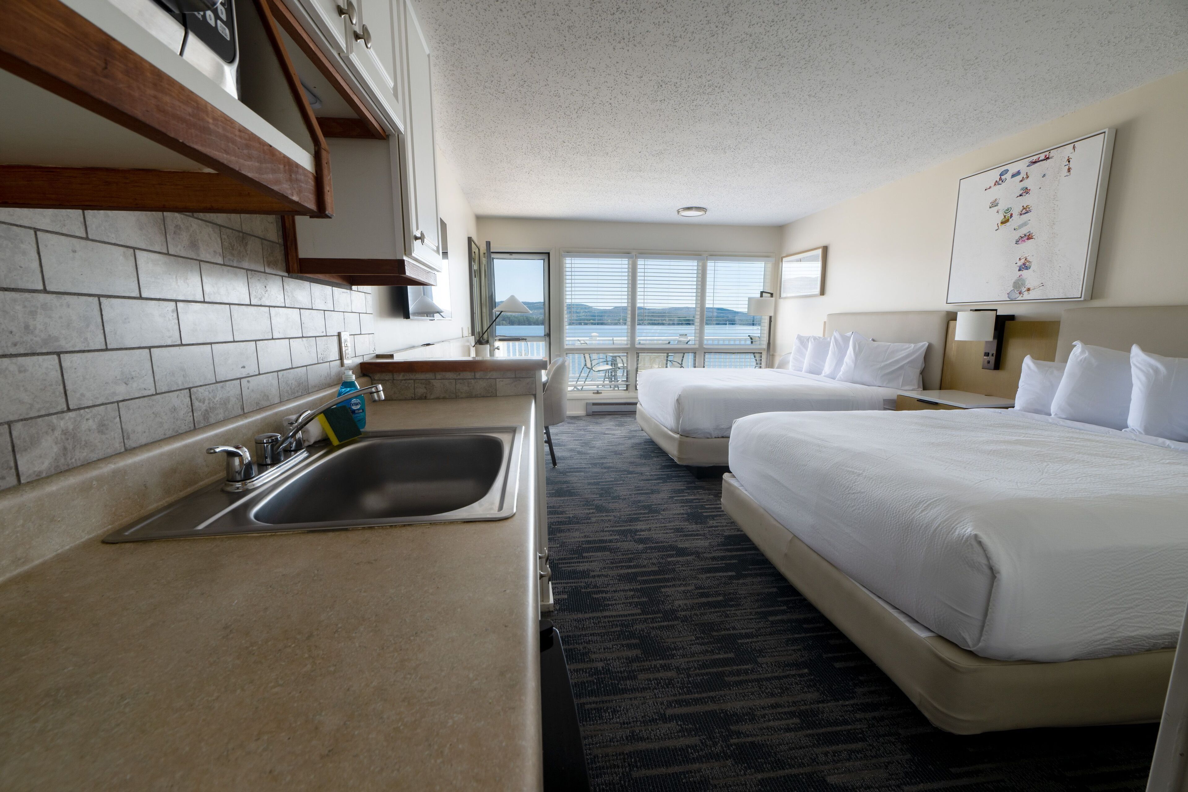 A hotel room with a kitchen area, two beds, a large window with a view, and a mounted artwork on the wall.