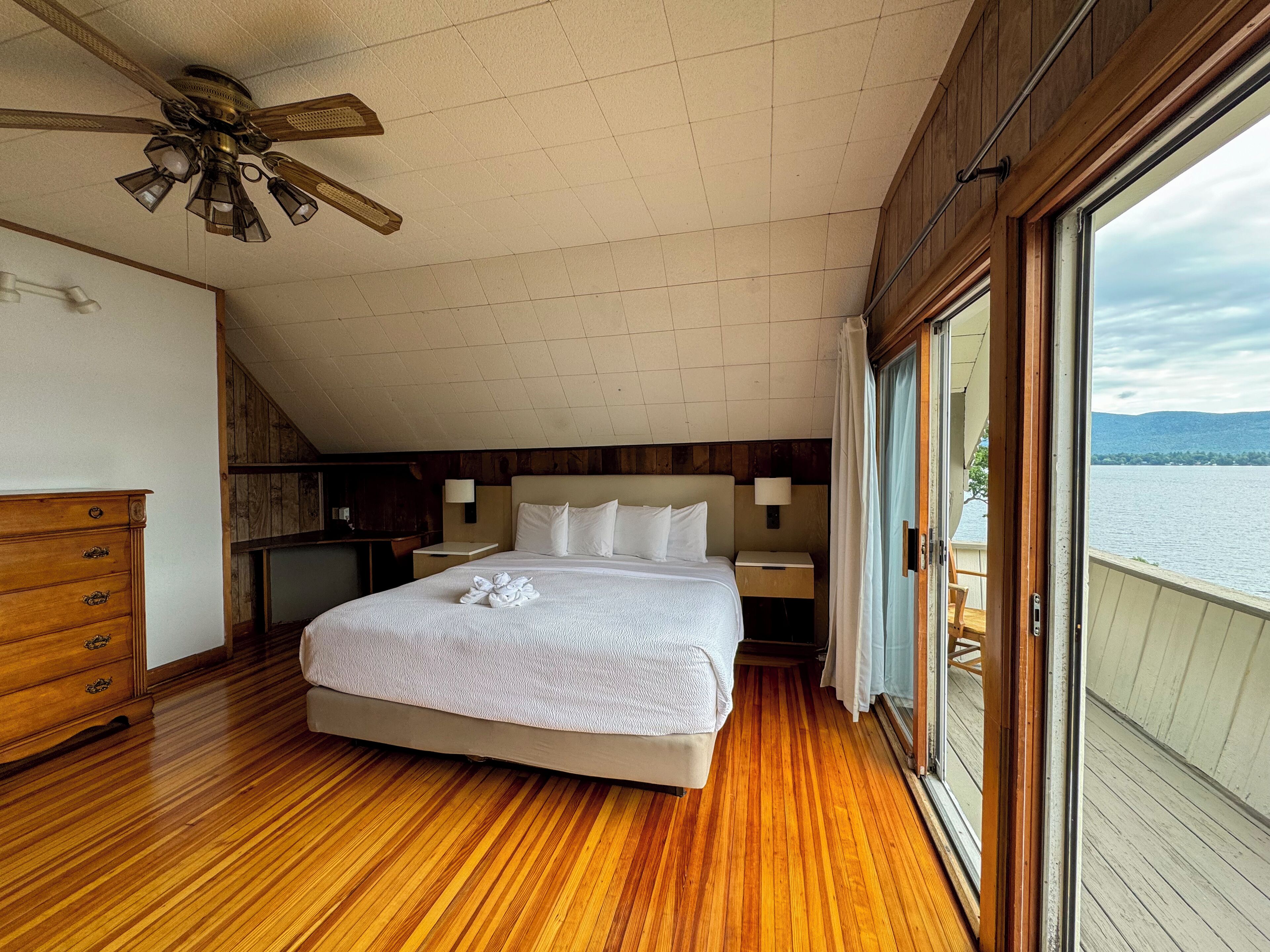 A bedroom with a large bed, wooden flooring, a ceiling fan, and a view of a lake.