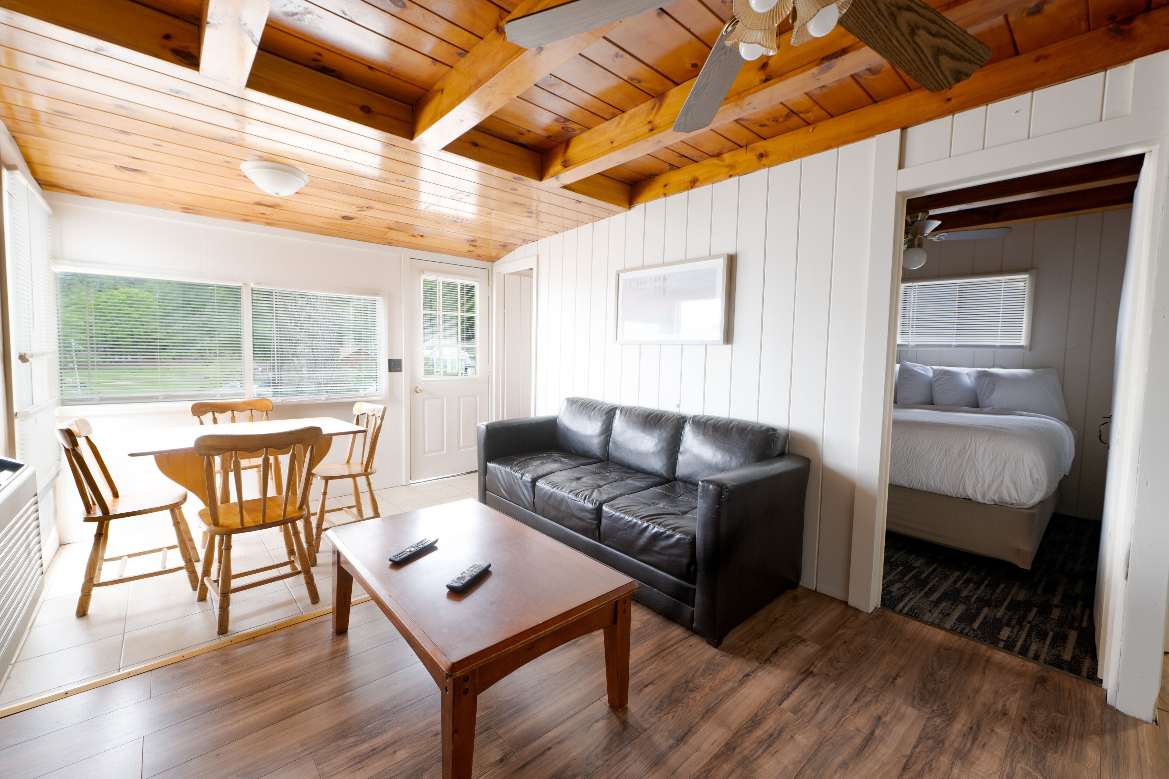 A cozy room with a wooden ceiling, a black leather sofa, a wooden dining table with chairs, a white bed, and a window with blinds.