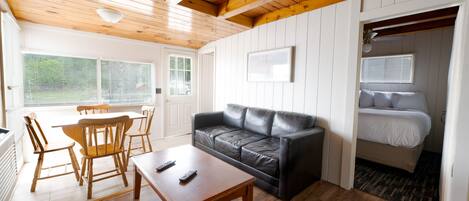 A cozy room with a wooden ceiling, a black leather sofa, a wooden dining table with chairs, a white bed, and a window with blinds.
