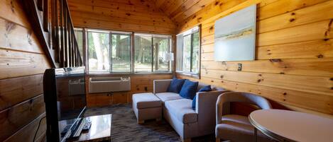 Un interior de cabaña acogedor con techo de madera, un conjunto de sofás, una mesa de comedor y ventanas grandes.