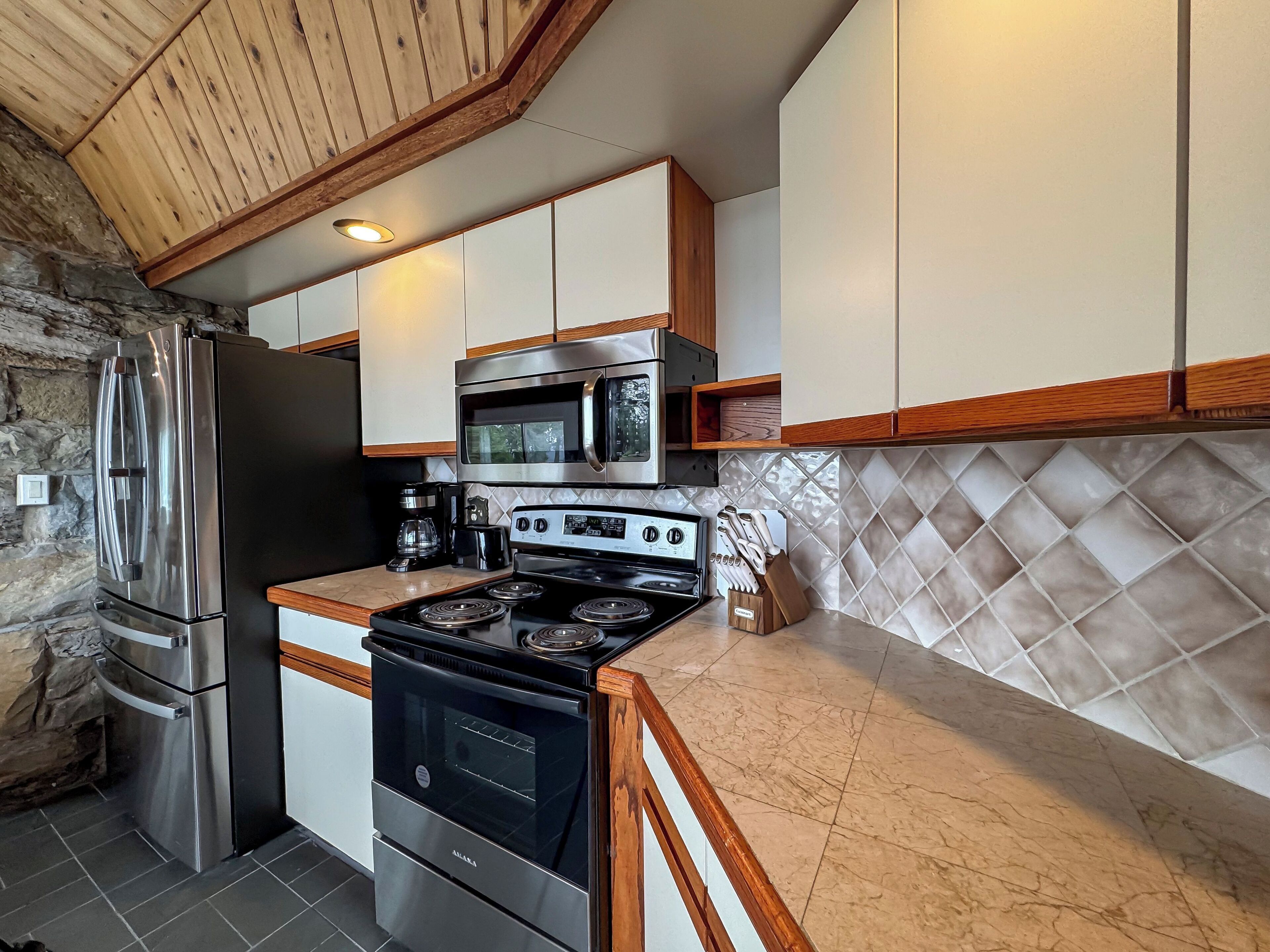 A kitchen with a stone wall, stainless steel appliances, wooden cabinets, and a tiled backsplash.