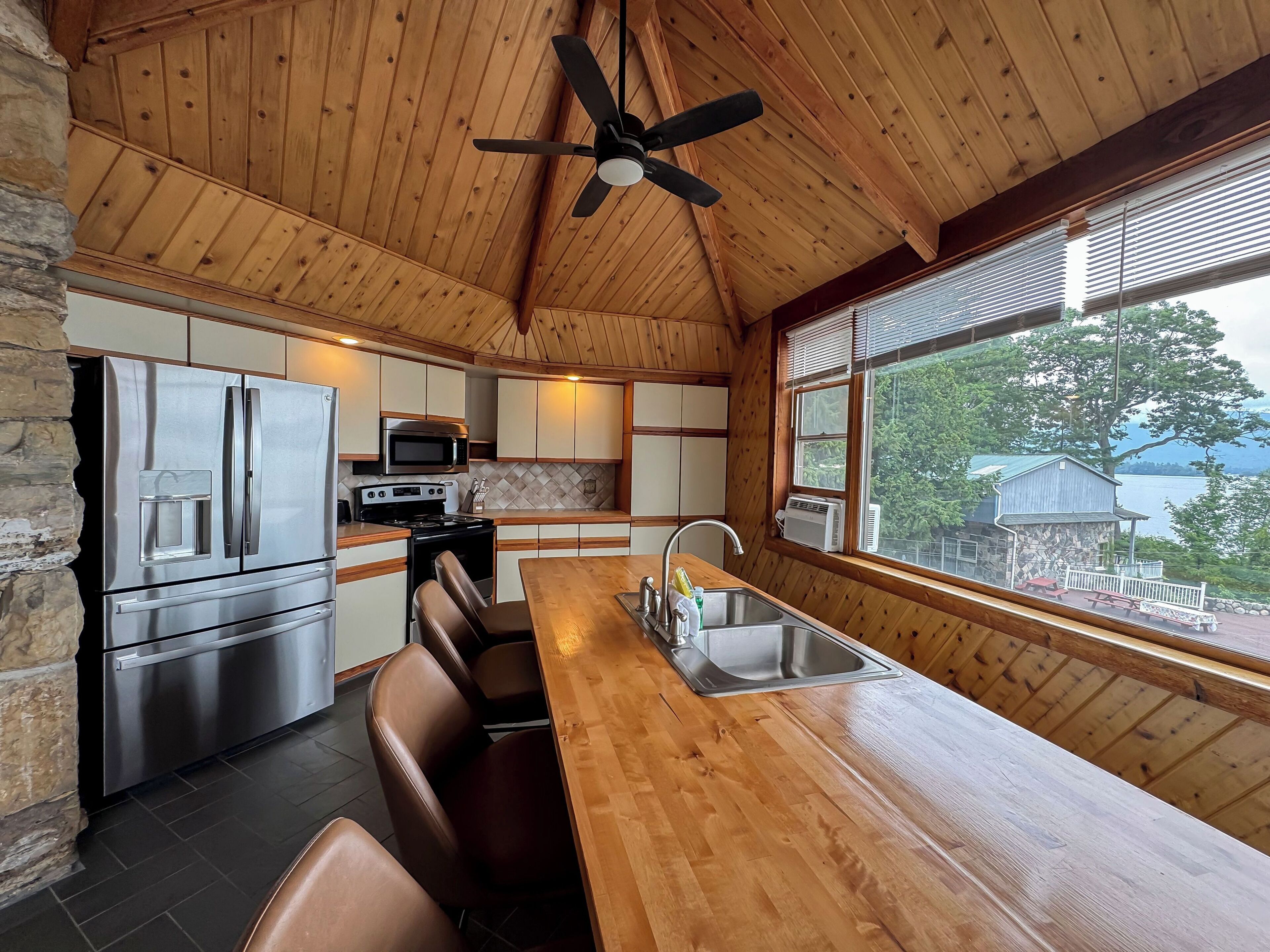 A kitchen with a wooden ceiling, stone wall, stainless steel appliances, and a large window overlooking a lake.