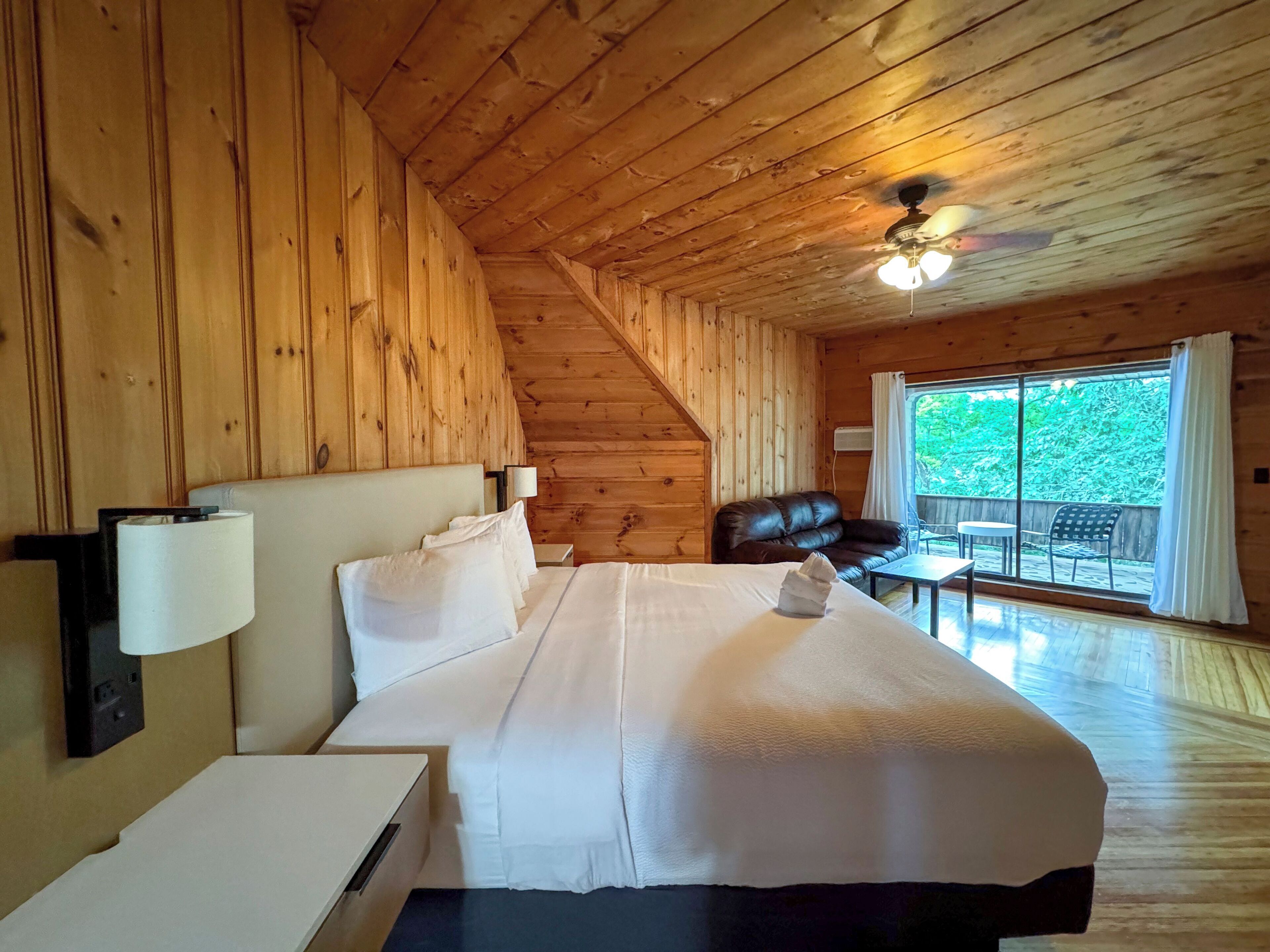 Een houten slaapkamer met een bed, een nachtkastje, een leren bank en uitzicht op de natuur door een groot raam.