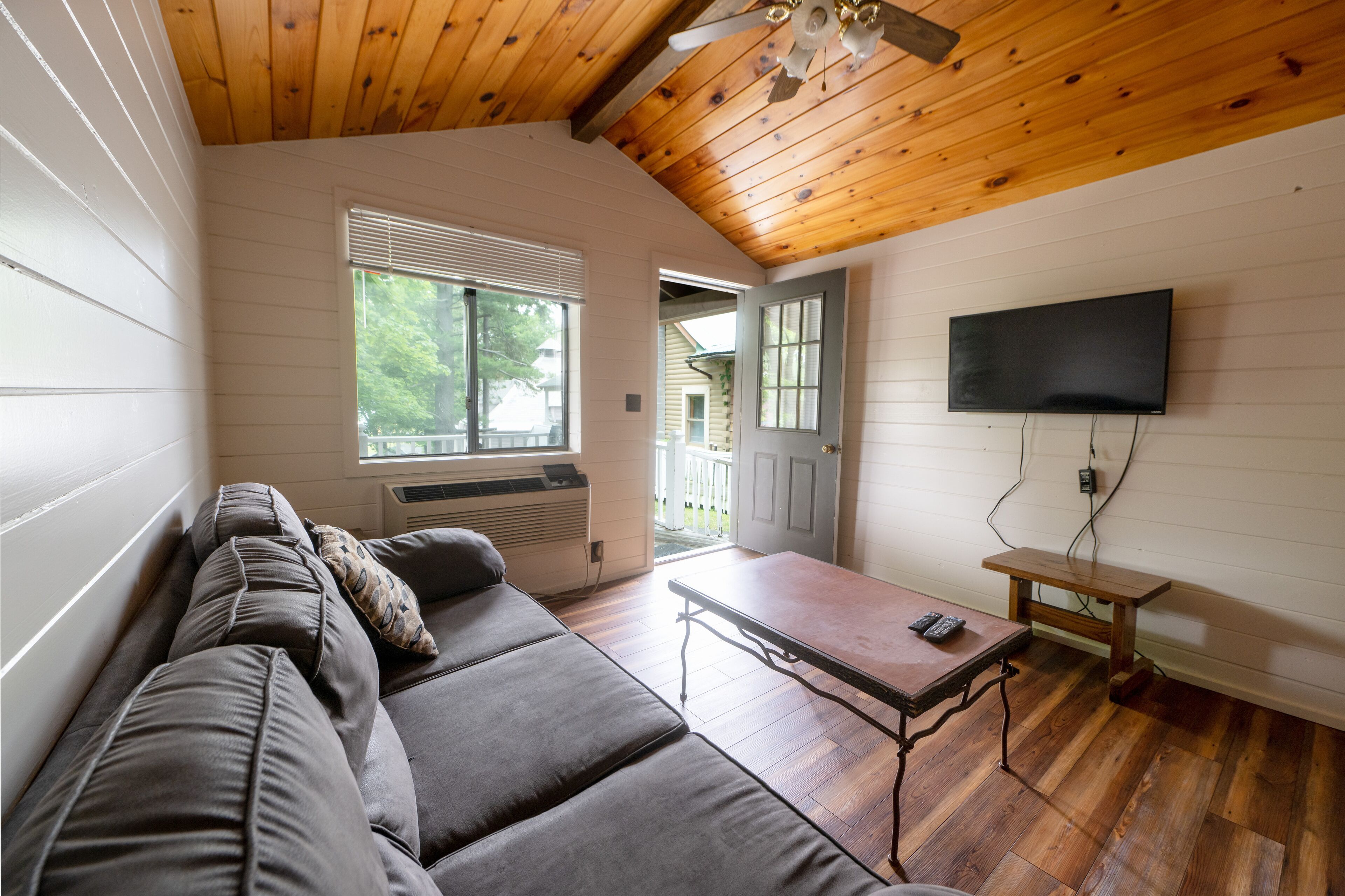 A room with a wooden ceiling, a sofa, a table, and a TV mounted on the wall.
