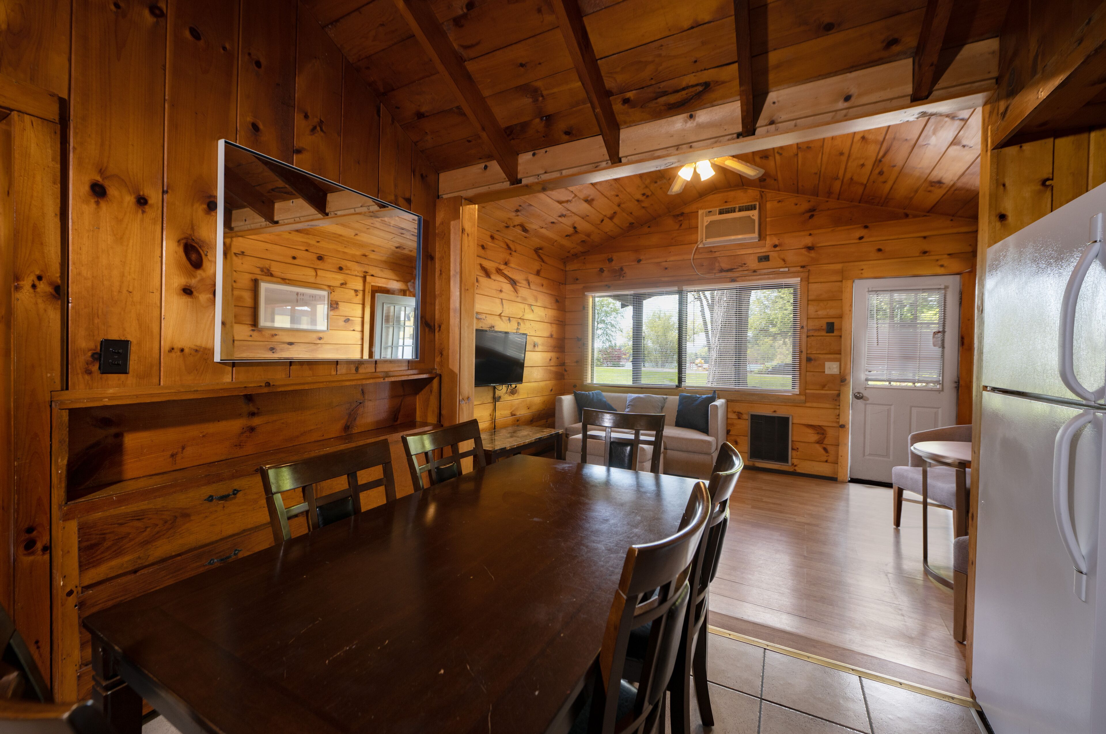 A wooden cabin interior with a dining table, chairs, a refrigerator, and a window.