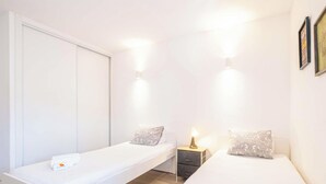 A small, well-lit bedroom with two beds, a bedside table with a lamp, and a wardrobe.