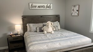 Un dormitorio con cabecera de madera, una cama grande con ropa de cama estampada, un peluche de elefante suave, una mesita de noche con una lámpara, y un letrero motivacional en la pared.