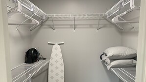 Un vestidor pequeño con estantes de metal blanco, un leña, una cesta de ropa y una cama con almohadas.