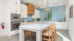 A modern kitchen with white cabinetry, a large island with chairs, stainless steel appliances, and a blue-tiled backsplash.