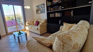 A living room with a sofa, a coffee table, and a bookshelf. There is a balcony with a purple chair and a TV mounted on the wall. - Detached Property with garden and a view on the Sea near Palermo, Sicily (Altavilla Milicia, Torre Colonna-Sperone)