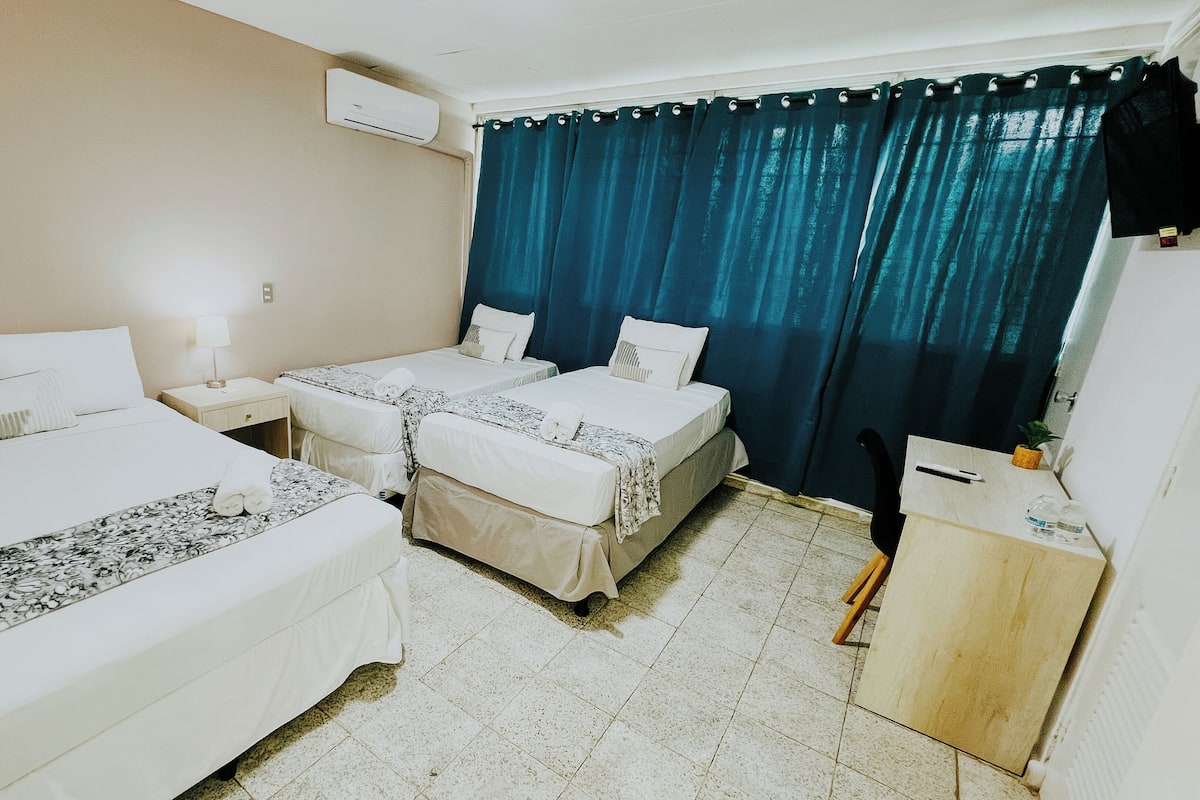 Habitación de hotel con dos camas, una mesita de noche con lámpara, un armario pequeño y una ventana con cortinas azules.